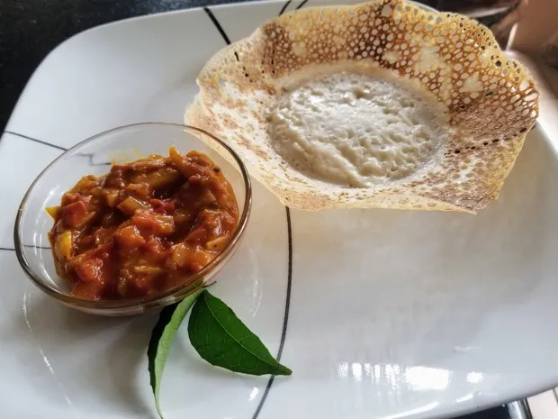A lacy appam with a glass bowl of tomato-and-vegetable curry and two fresh curry leaves