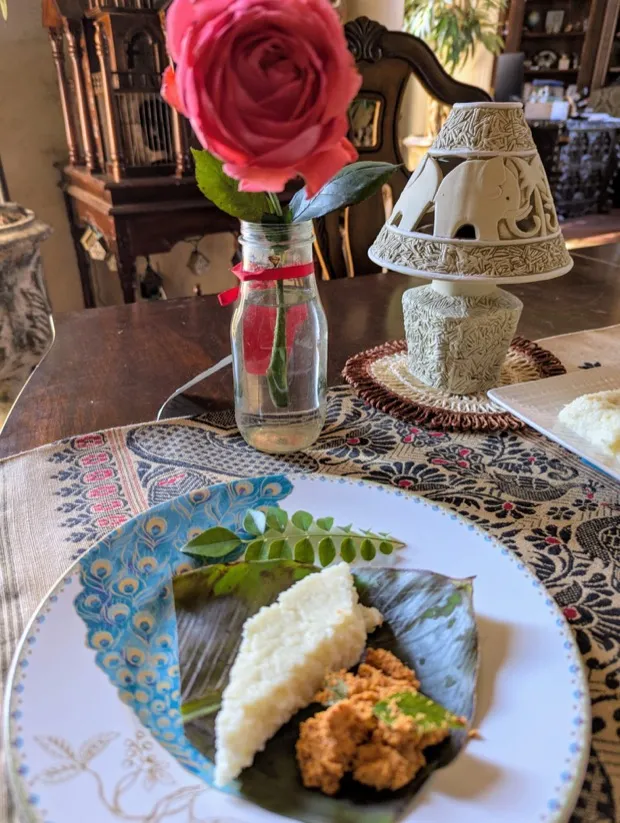 A rice cake and a spoonful of chutney podi served on a banana leaf, on a table laid with a rose and a carved lamp