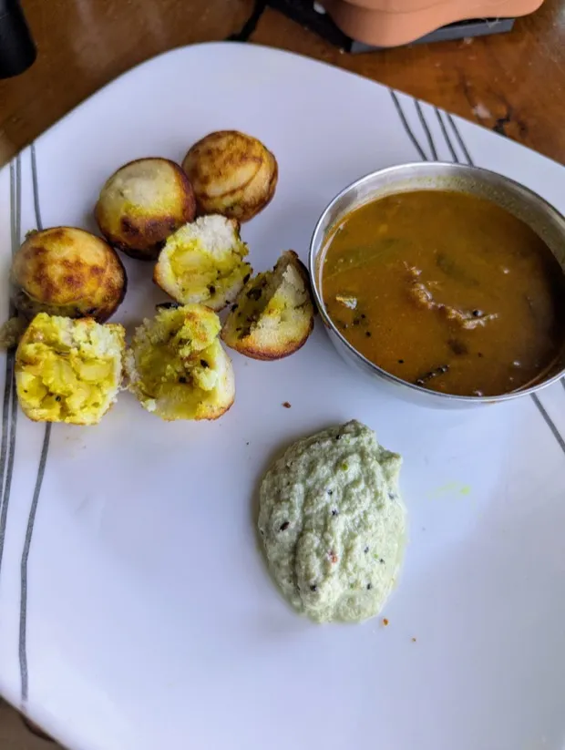 Rice-batter paniyaram broken open to show a spiced potato filling, served with sambar and coconut chutney