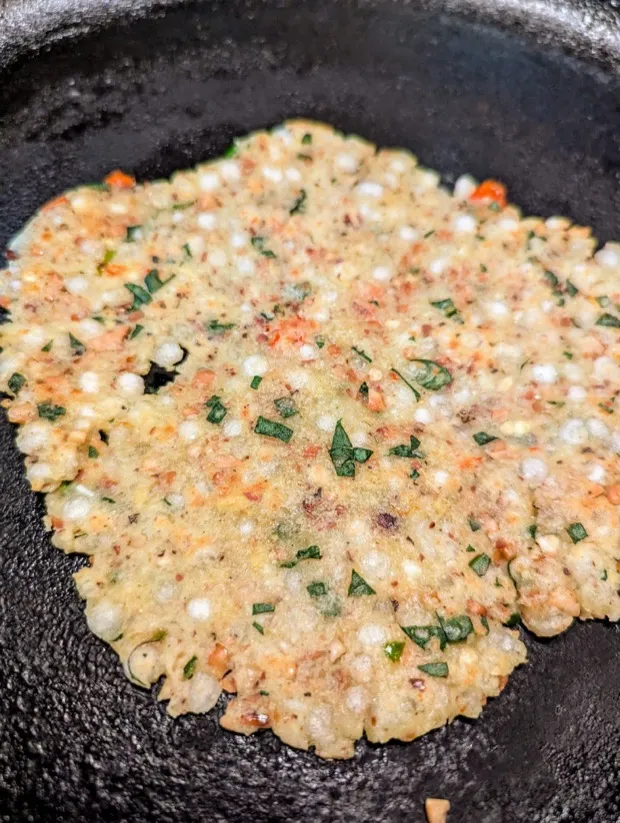 A tapioca-pearl thalipeeth studded with crushed peanuts and coriander, cooking on a cast-iron pan