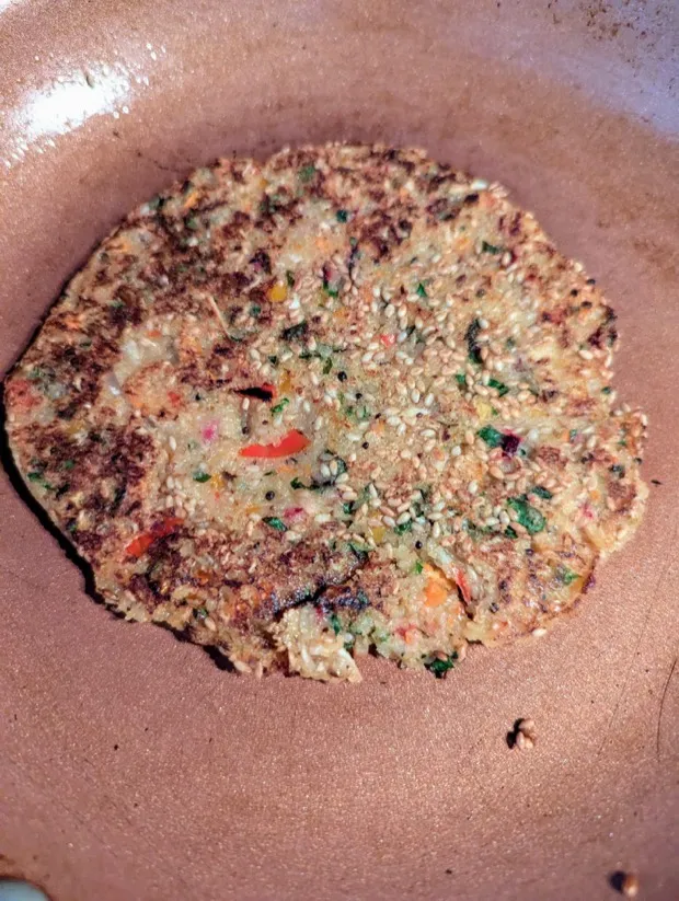 A sesame-crusted vegetable thalipeeth browning in a pan, flecked with coriander, carrot and chilli