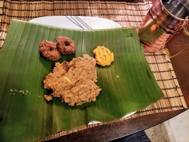 Two medu vadai and a mound of sweet pongal served on a banana leaf, with a copper tumbler alongside