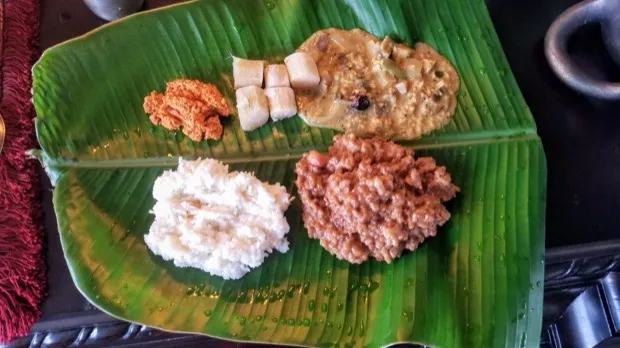 A banana-leaf plate set with chutney podi, rice-cake pieces in a mild curry, fresh grated coconut and a groundnut-jaggery crumble