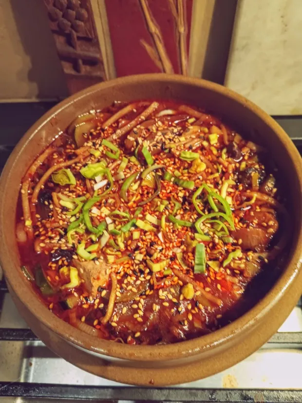 A clay pot of noodle soup in a red broth, finished with sesame seeds and sliced spring onion