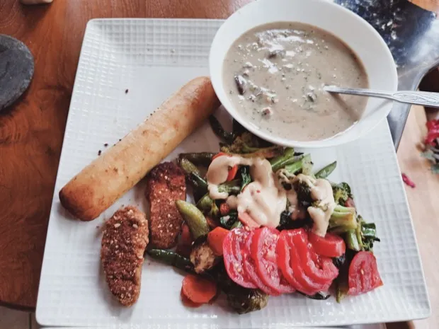 A breadstick and two crumb-coated cutlets with roasted vegetables, sliced heirloom tomato and a bowl of creamy mushroom soup