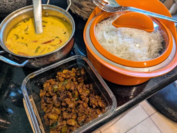 A pan of coconut curry beside a container of freshly steamed string hoppers and a box of dark spiced stir-fry