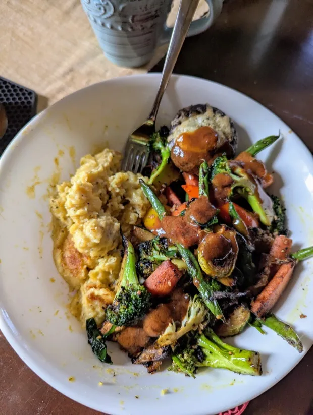 A bowl of mash with roasted broccoli, carrots, green beans and brussels sprouts, cheese-topped mushroom caps and a drizzle of peanut sauce