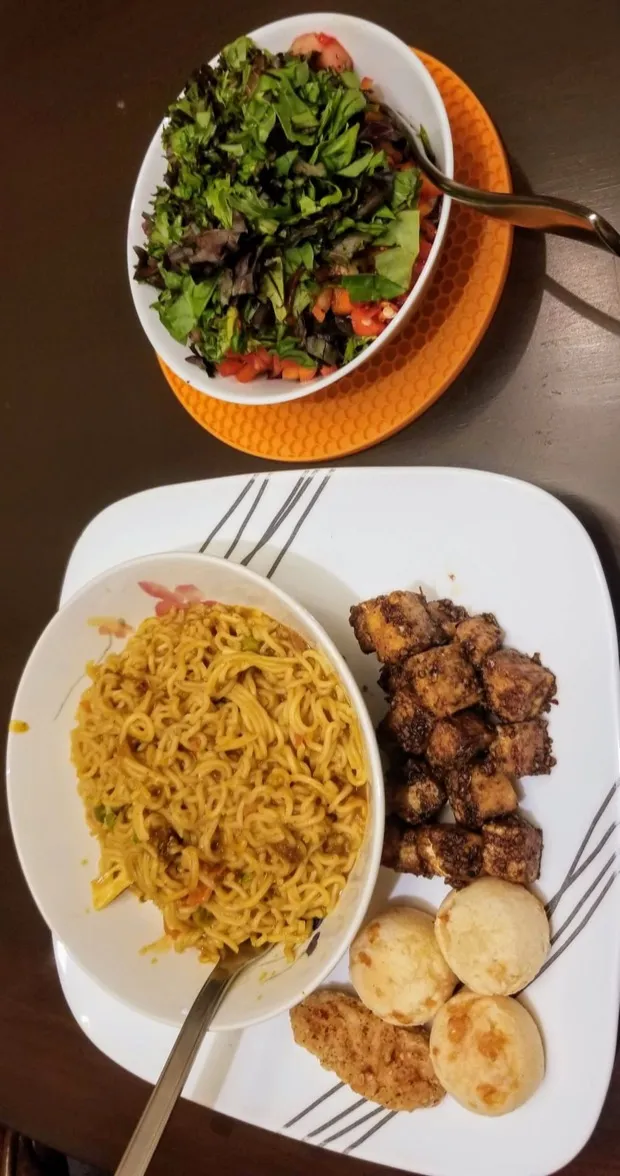 A bowl of vegetable noodles with spiced tofu cubes, soft baked buns and a cutlet, with a bowl of leafy salad above