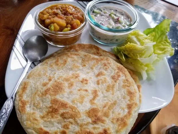 A flaky parotta with a bowl of potato curry, a bowl of cucumber raita and crisp lettuce leaves