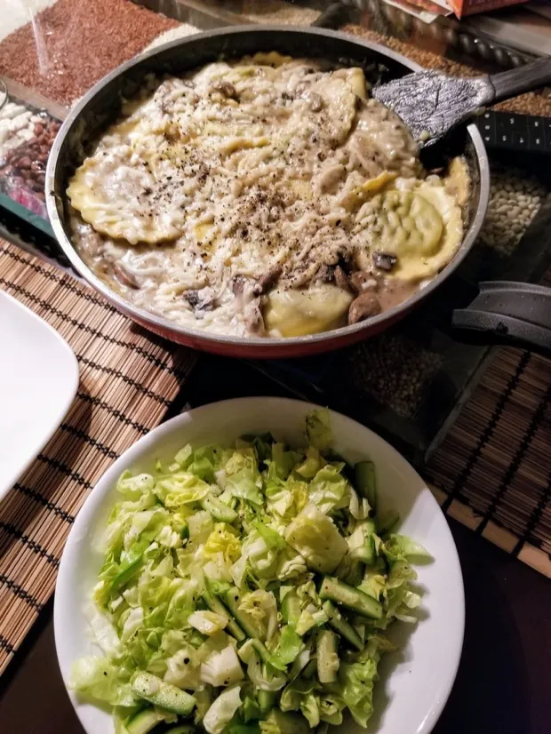 A pan of ravioli in a creamy mushroom sauce with cracked pepper, and a bowl of chopped lettuce and cucumber salad