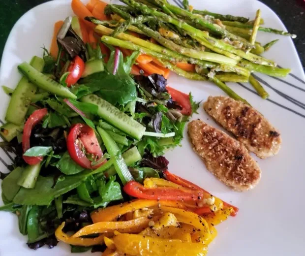 A leaf salad with cucumber and tomato beside roasted asparagus, sautéed yellow and red peppers and two crumbed cutlets
