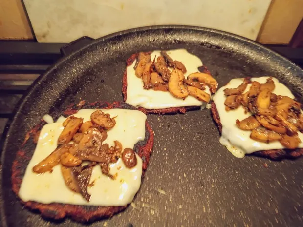 Beetroot patties topped with melted cheese and sautéed mushrooms on a griddle