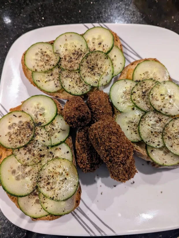 Three slices of toast fanned with thin cucumber rounds and cracked pepper, with crumb-coated cutlets between them