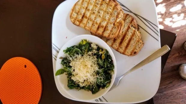 Two grilled slices of seeded sourdough with a bowl of sautéed greens under grated cheese